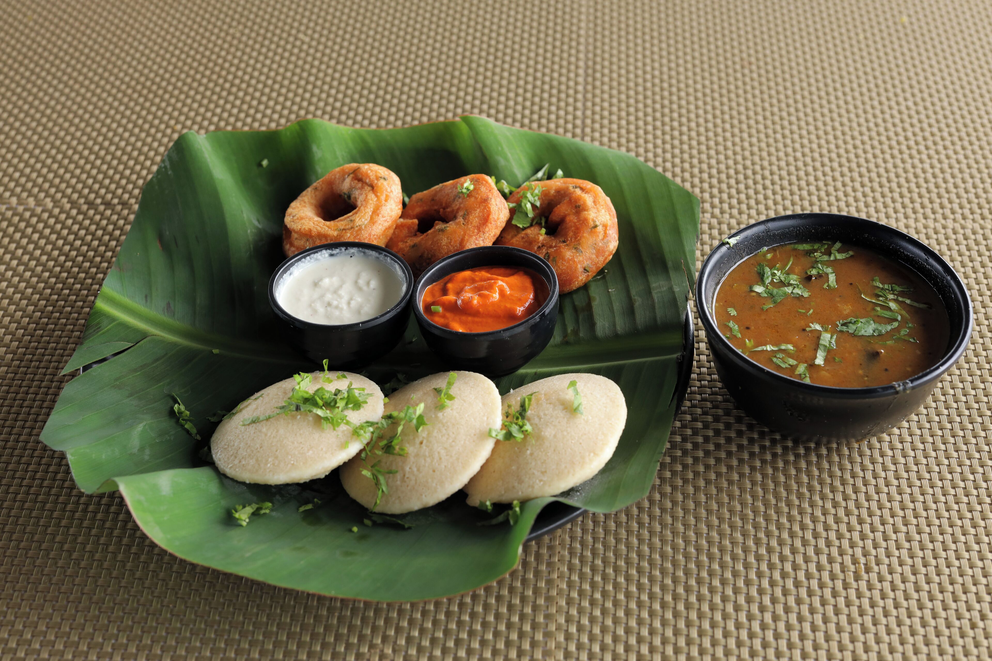 Une assiette de nourriture comprenant des idlis, des vadas et deux sortes de chutney, le tout servi sur une feuille de bananier.