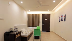 Habitación de hotel con cama, una silla verde, un escritorio con hervidor y botellas de agua, y una puerta con un cartel.