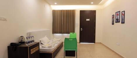 Habitación de hotel con cama, una silla verde, un escritorio con hervidor y botellas de agua, y una puerta con un cartel.