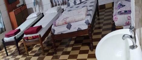 Habitación de hotel con piso a cuadros, cama con ropa de cama floral, mesita de noche con cojín rojo, cabecera de madera, lavamanos blanco y espejo.