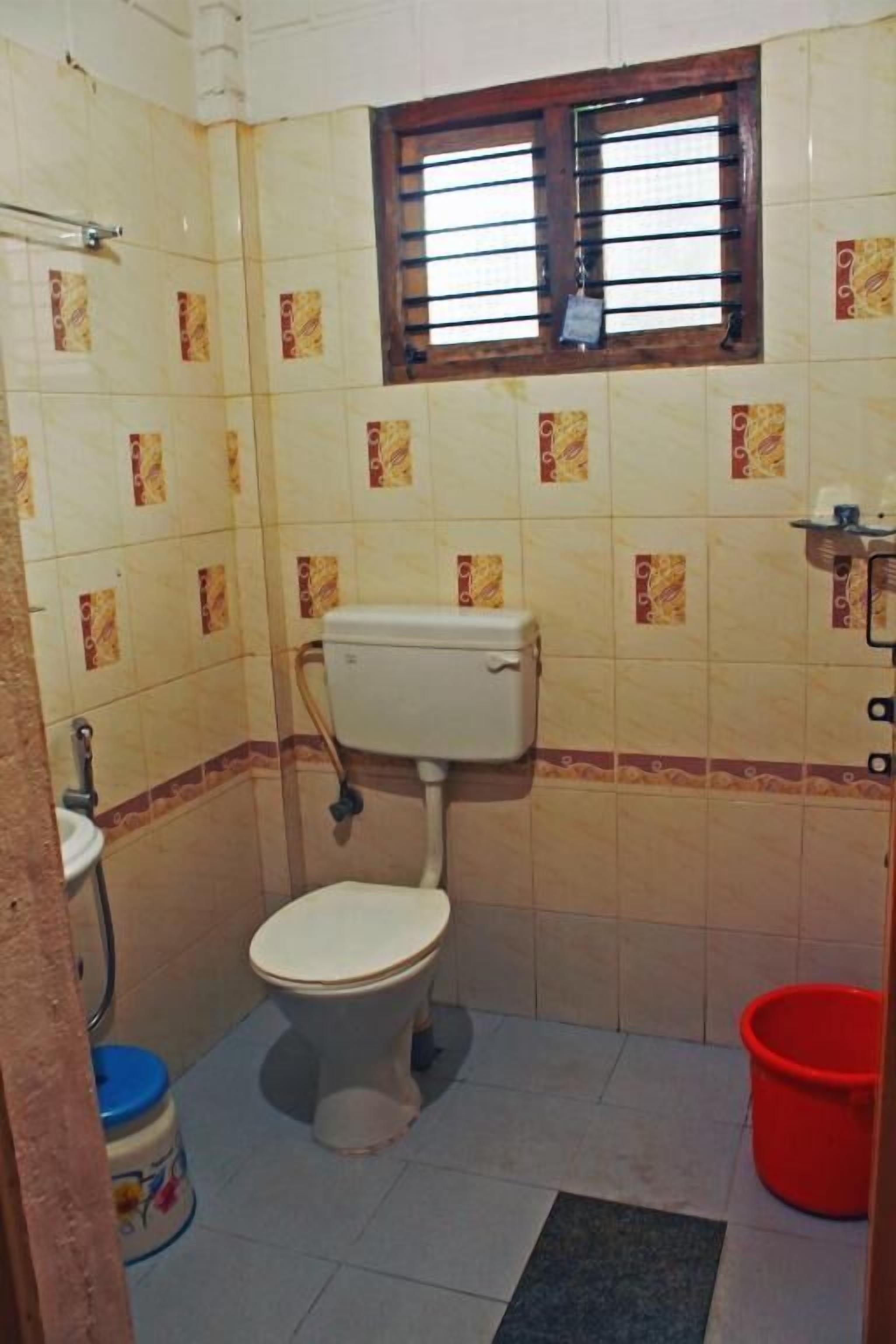 Un baño pequeño con un inodoro, una ventana con barrotes, un cubo rojo y una pared de azulejos con un diseño.