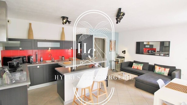 Una cocina moderna con armarios grises y un salpicadero rojo, una isla central con taburetes, y un sofá negro con cojines coloridos en una zona de estar.