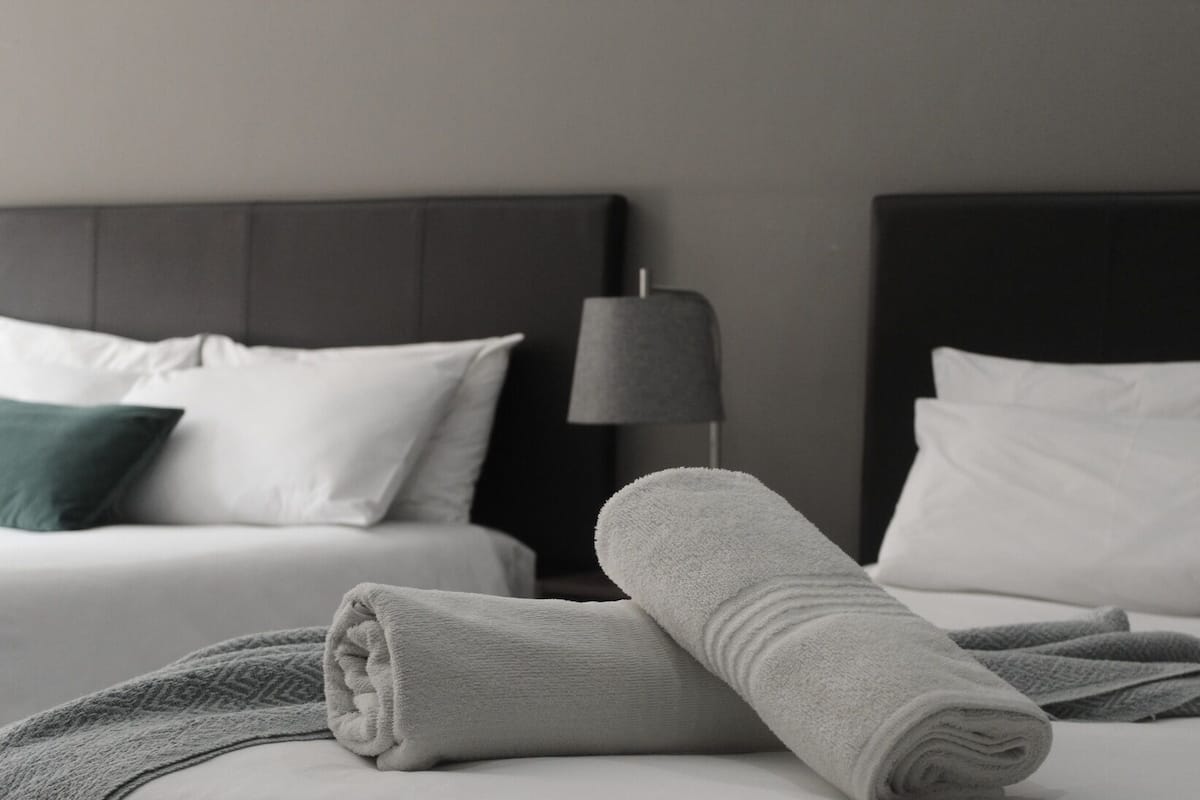 Cama bem arrumada com toalha enrolada, duas almofadas brancas e abajur escuro ao lado.