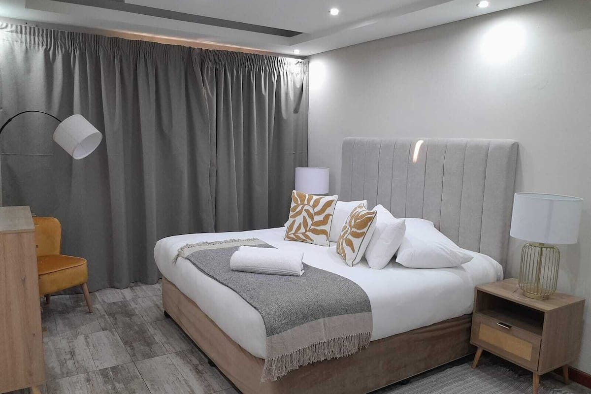 Quarto de hotel moderno com uma cama grande, uma mesa de cabeceira, uma luminária e uma cadeira.