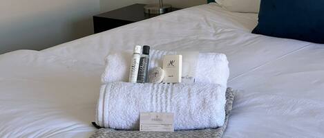A neatly made bed with a white comforter, a grey throw blanket, and a set of toiletries including lotion, shampoo, and a hair cap.