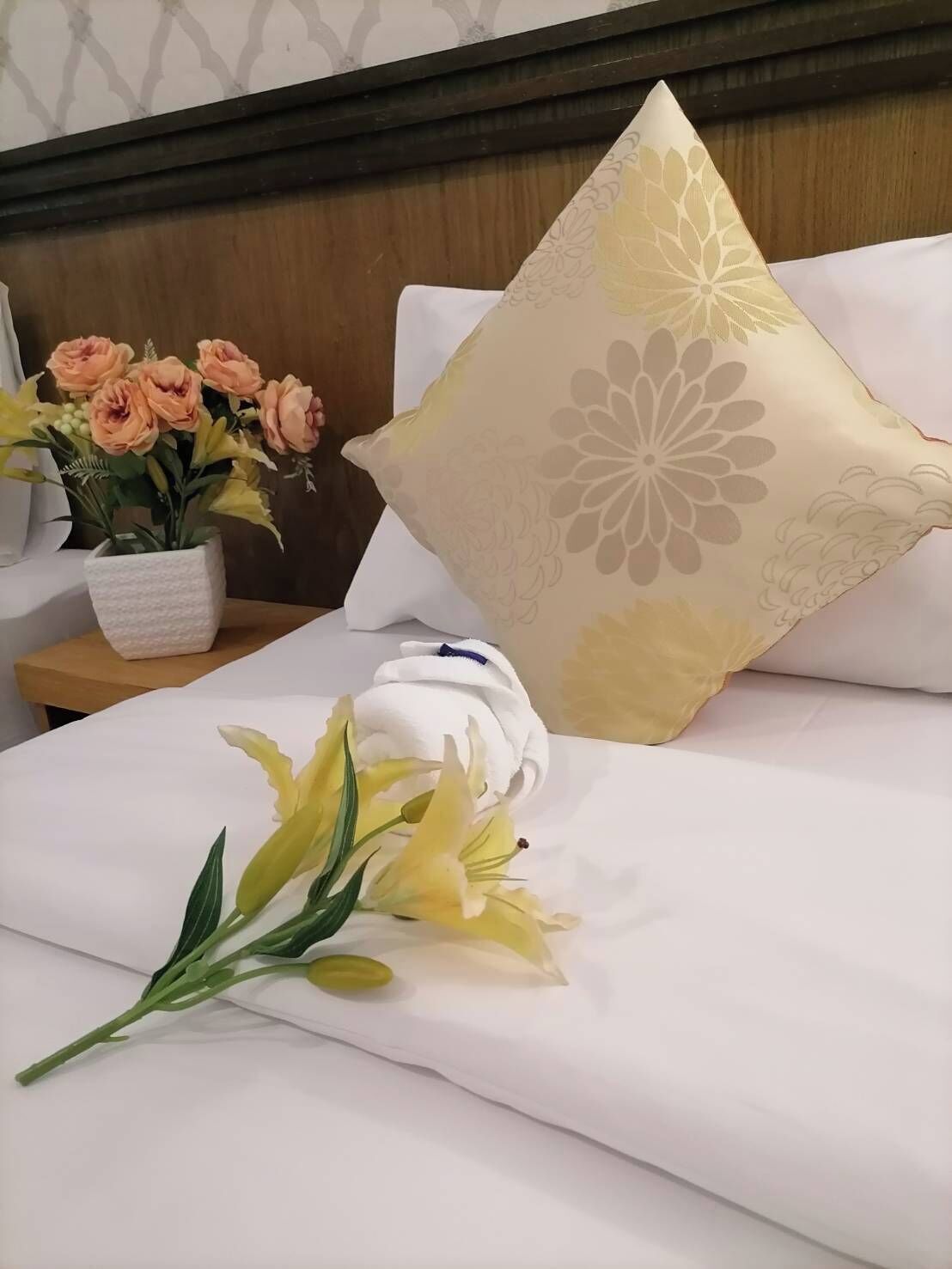 Een opgemaakt bed met witte lakens, een decoratief kussen met bloemenmotief en een bloemenvaas op een nachtkastje.