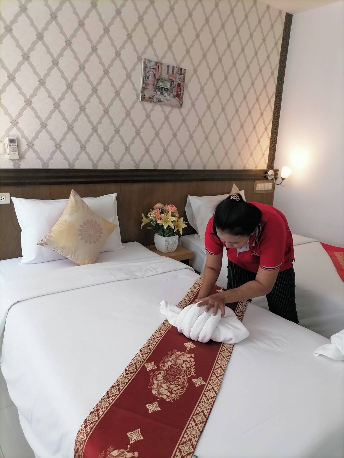 A neatly made bed with a decorative red runner, a floral arrangement on a bedside table, and a patterned wall.