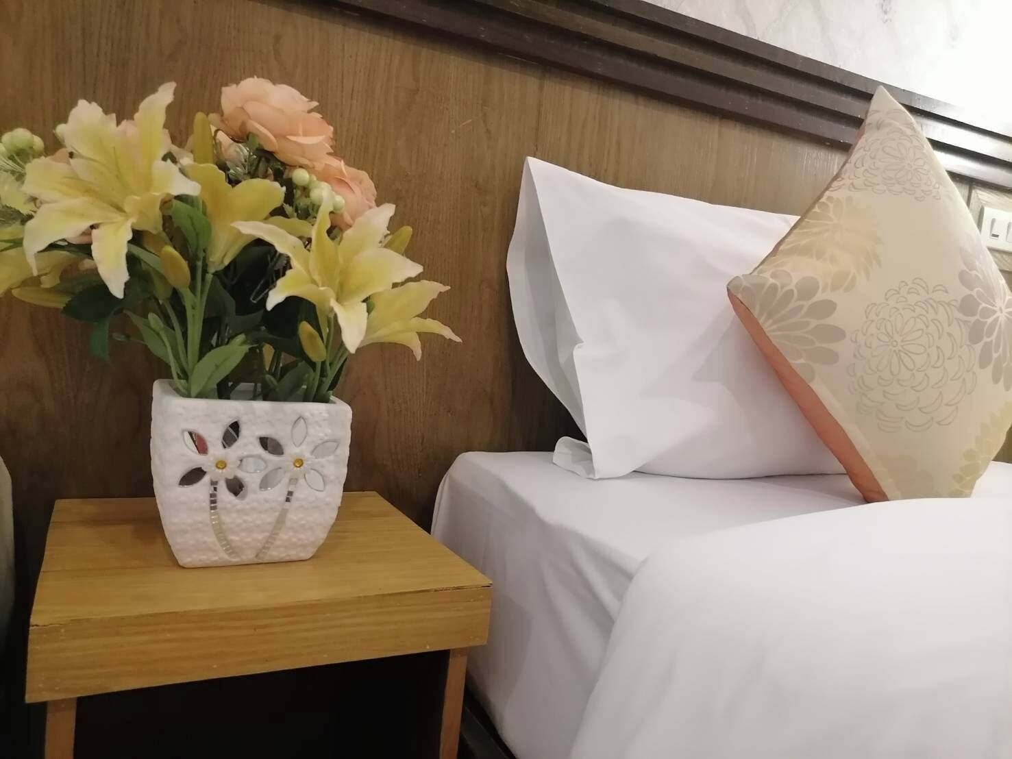 A bed with white bedding and a decorative pillow with a floral pattern. A small wooden nightstand holds a white vase with a floral design containing yellow and peach flowers.