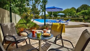 An outdoor patio with a table set for two, surrounded by chairs, a pool, and lounge chairs in the background. - Nyktelia Holiday Villa (Lefkada)