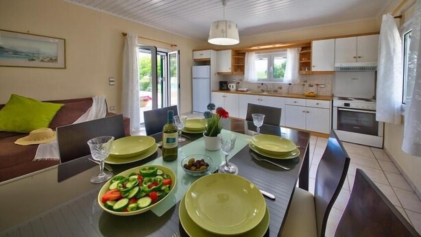 A modern kitchen with white cabinets, a dining table set for four, and a view of the outdoors through the window. - Nyktelia Holiday Villa (Lefkada)