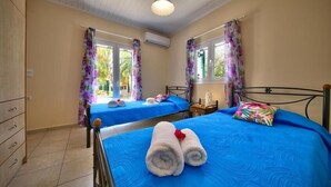 A hotel room with two beds, each with a blue bedspread and white towels. There is a wooden wardrobe, a small table with a lamp, and a window with floral curtains. - Nyktelia Holiday Villa (Lefkada)