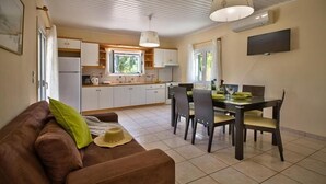 A cozy living room with a brown sofa, a dining table with chairs, and a kitchen area with white cabinets and a window. - Nyktelia Holiday Villa (Lefkada)