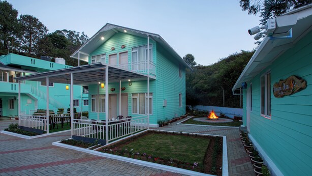 A turquoise two-story building with a covered patio area, a small fire pit, and a row of potted plants along the side. - Blossom Inn (Coonoor)