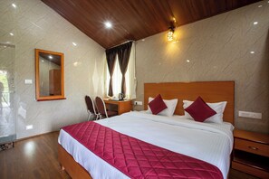 A hotel room with a large bed, wooden headboard, a desk with a chair, a mirror, and a window with curtains.