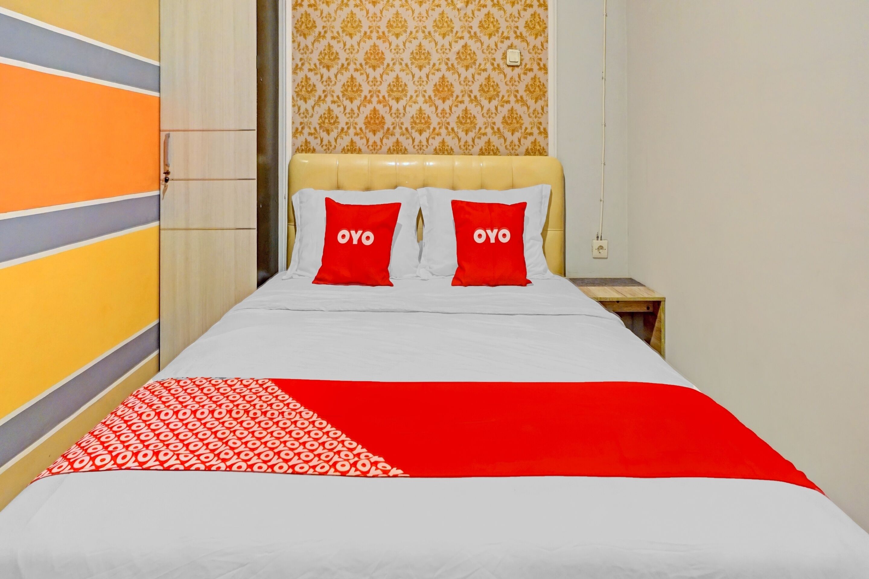 A hotel room with a bed, two red pillows with the word "OYO" on them, a patterned red and white throw blanket, and a wooden nightstand.