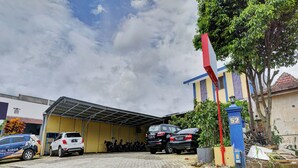 A parking area with several cars, a covered structure, and a building with a blue and yellow facade. - Hotel O Btc Guest House Syariah (Malang)