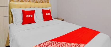 A neatly made bed with a white and red patterned bedspread, two red pillows with "OYO" text, and a wooden nightstand beside it.