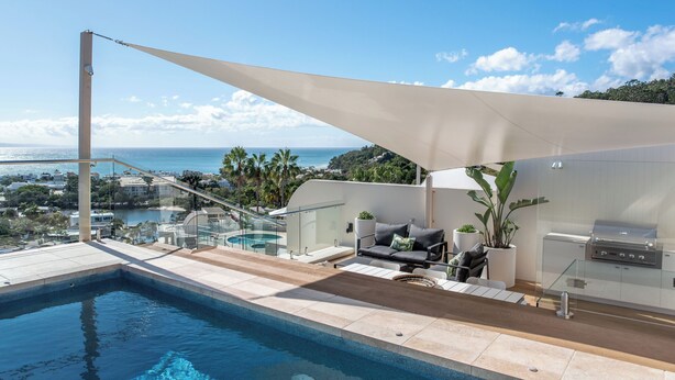 A modern outdoor pool area with a view of the ocean, a barbecue, and a seating area with cushions and plants.