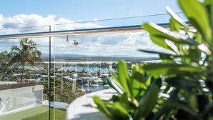 A balcony with a glass railing overlooking a coastal town, a clear sky, and some greenery in the foreground.