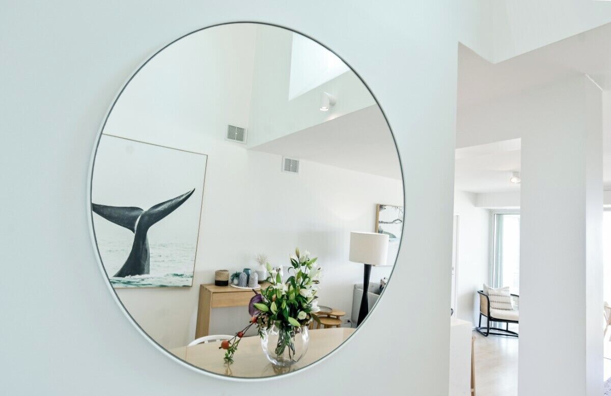 A round mirror on a wall with a whale tail painting, a vase with flowers, and a desk with a lamp.