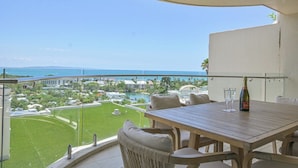 A balcony with a wooden table, two chairs, and a bottle of champagne. A glass is also visible on the table. The balcony overlooks a green lawn and a body of water with buildings in the background.