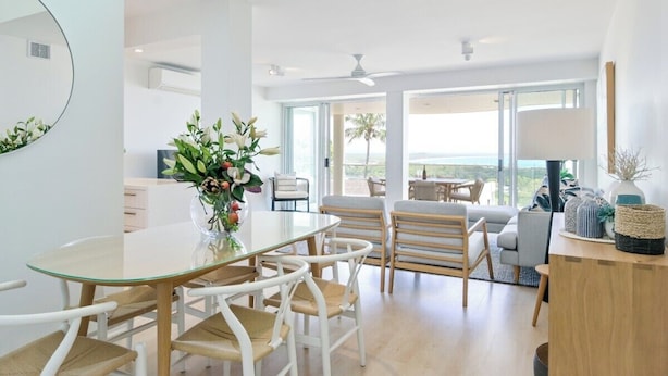 A modern living room with a glass dining table, white chairs, and a view of the ocean through large windows.