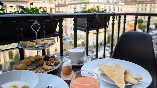 A breakfast setup on a balcony with a city view, including a plate of pastries, a bowl of cereal, a glass of orange juice, and a cup of coffee. - Mastropasqua BB (Naples)