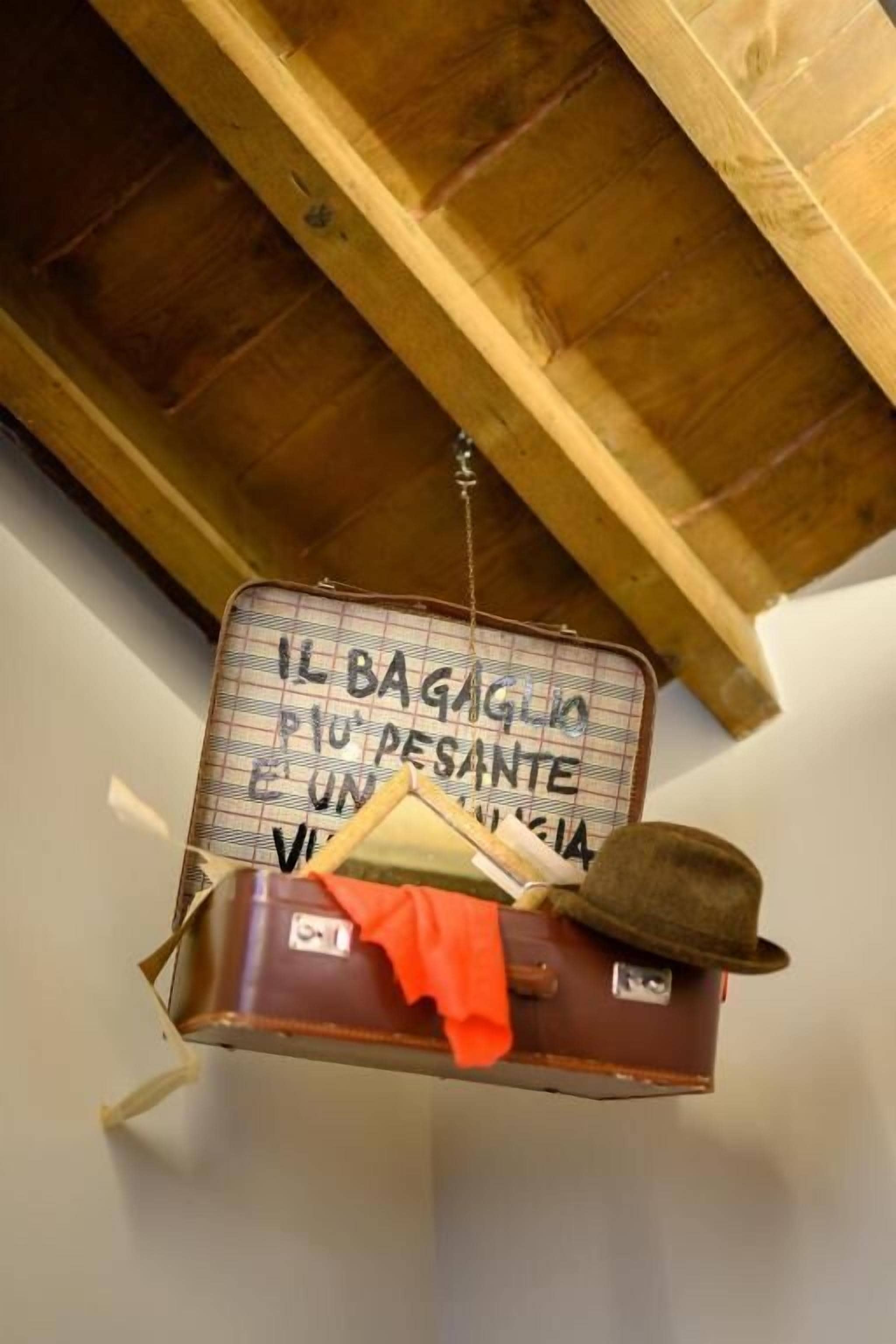 A suitcase hanging from a wooden beam with a handwritten sign that reads "IL BAGAGLIO PIÙ PESANTE E UN PIÙ SANTESSE."