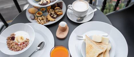 Una mesa con alimentos para el desayuno, incluyendo tostadas, yogur, un vaso de jugo de naranja y una variedad de pasteles.