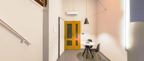A narrow hallway with a staircase on the left, a yellow door at the end, a round table with chairs, and a pendant light.