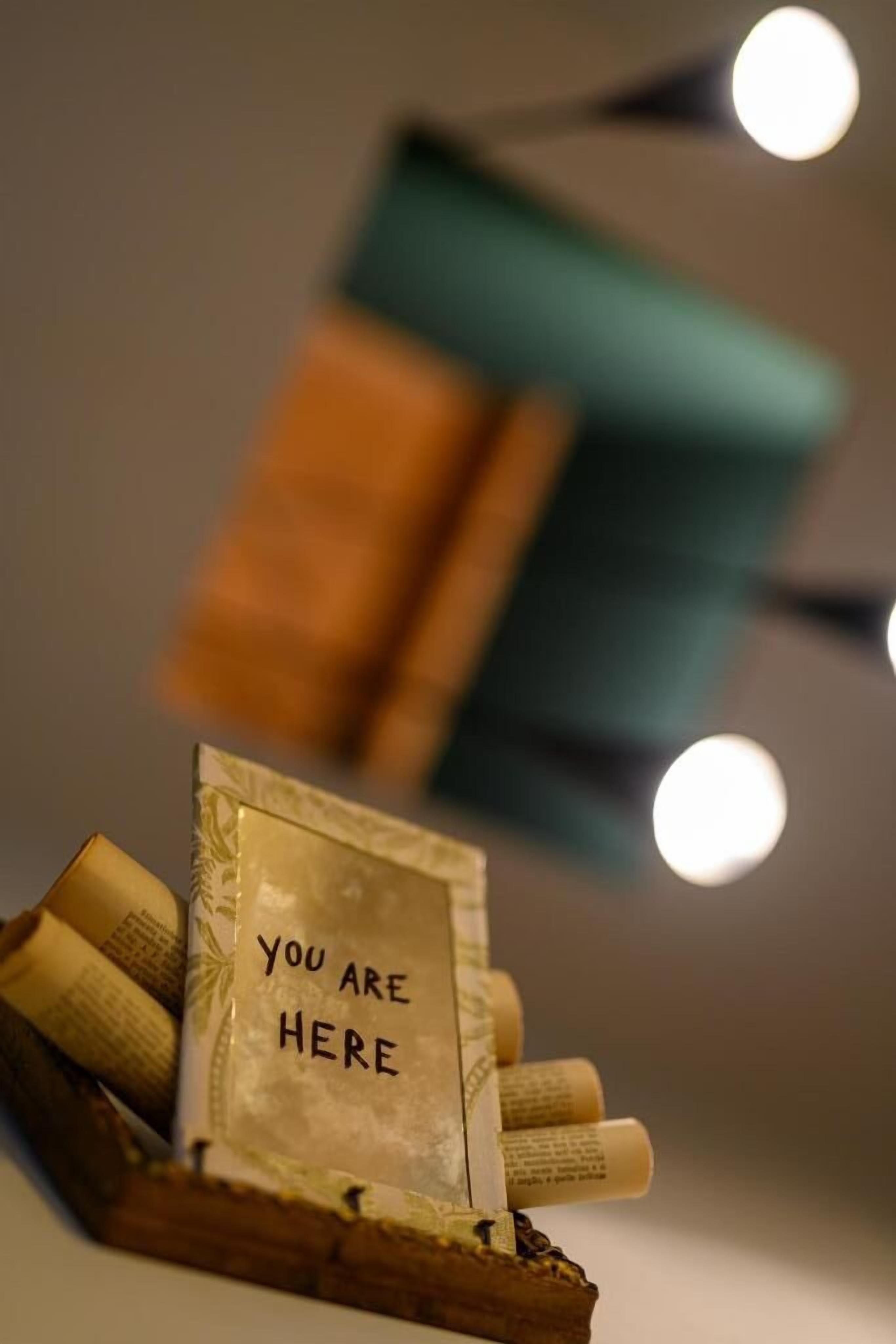 A framed message card with the text 'YOU ARE HERE' placed on a wooden surface with rolled-up papers.