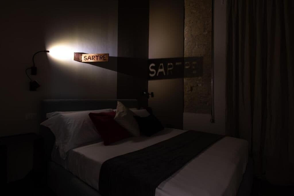 A dimly lit hotel room with a bed, a wall-mounted sign reading "SARTRE," and a wall with a textured surface.