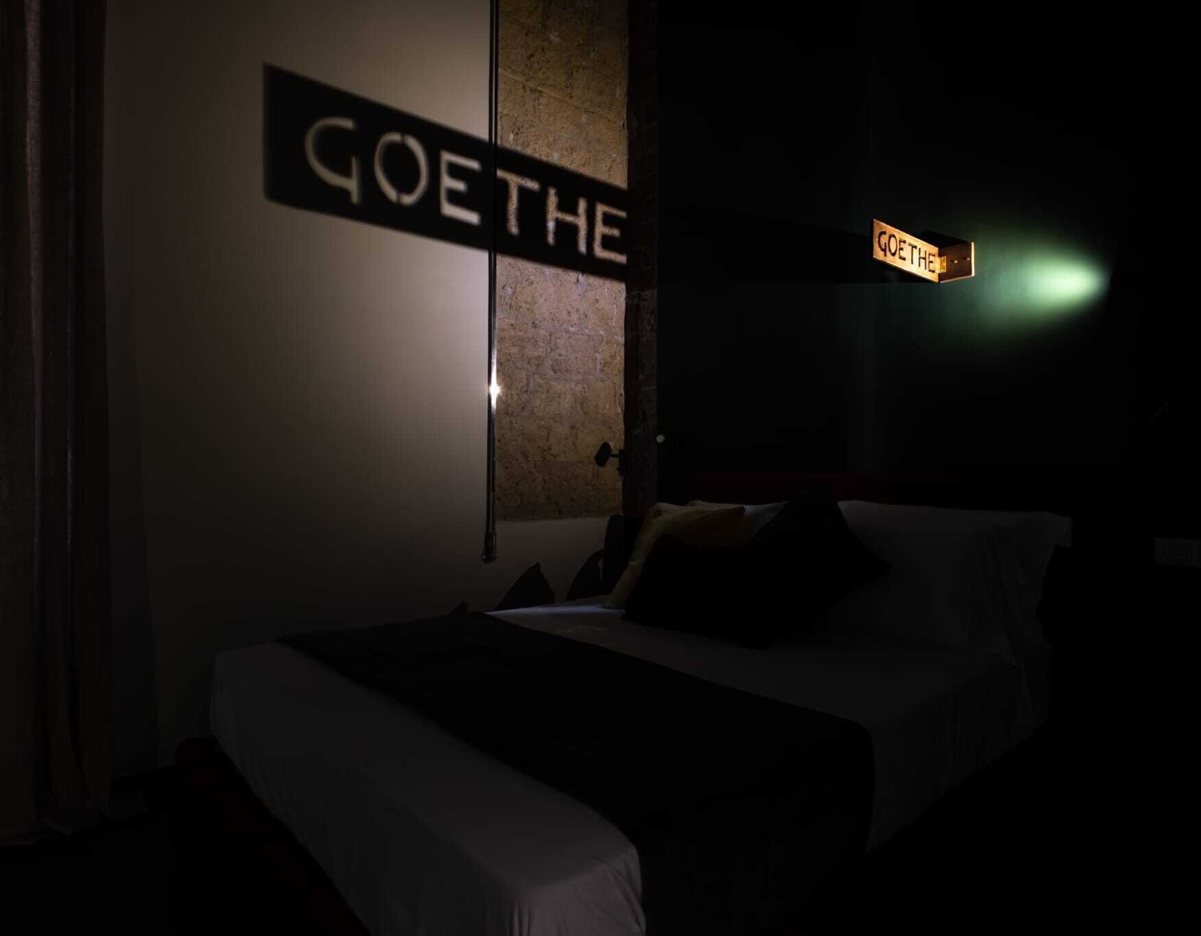 A dimly lit hotel room with a bed, a nightstand, and a sign that reads "GOETHE".