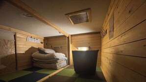Une petite pièce aux murs en bois, faiblement éclairée, avec un lit simple recouvert d’un matelas à carreaux verts et bruns. On y trouve une petite table sur laquelle est posée une lampe, ainsi qu’un radiateur fixé au mur.