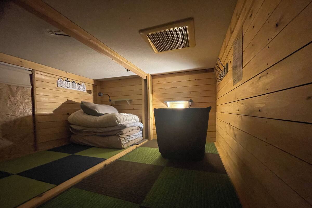 Una habitación pequeña, tenuemente iluminada, con paredes de madera y una cama individual con colcha verde y marrón a cuadros. Hay una mesita con una lámpara y un calentador fijado a la pared.