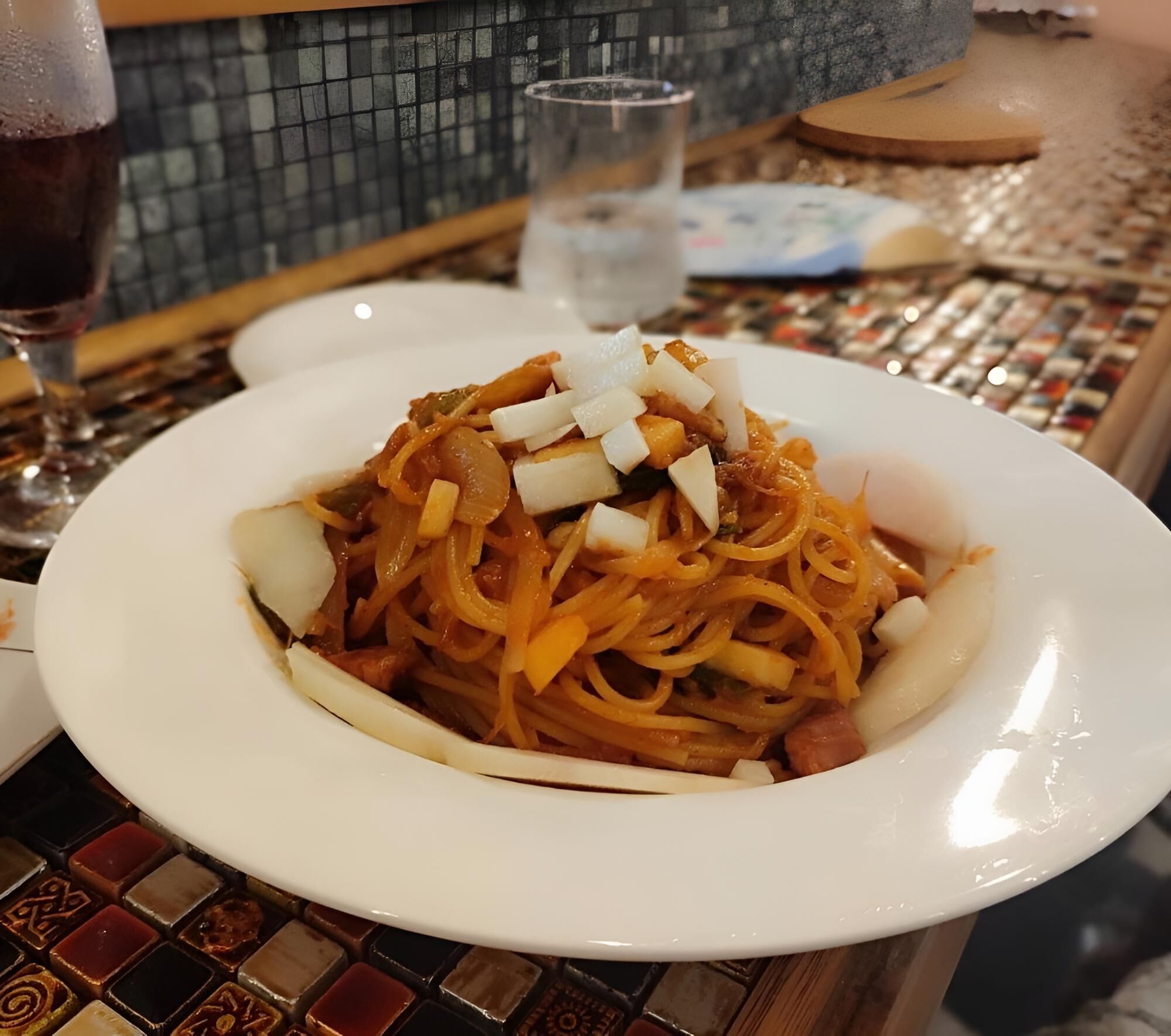 A plate of spaghetti with vegetables and meat, a glass of water, and a wine glass on a table with a tiled pattern.