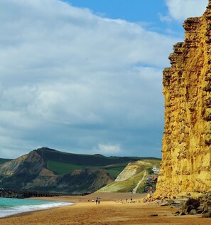 A coastal landscape with a sandy beach, cliffs, and a pier extending into the water. - Cottage close to Beach, Pub & Amenities (Charmouth)
