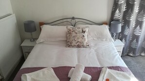 Una cama bien tendida con sábanas blancas, un cojín floral y toallas dispuestas sobre una colcha morada.