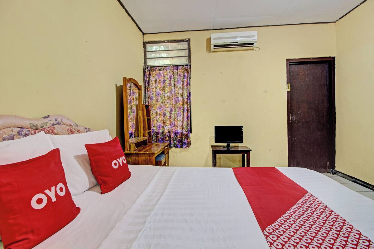 A hotel room with a bed, a wooden desk, a mirror, a television, and a door.