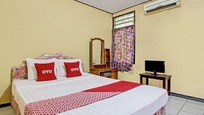 A neatly made bed with a patterned bedspread and two red pillows, a wooden nightstand with a television, a mirror, and a window with floral curtains. - Hotel O Sendang Asri (Wonogiri)