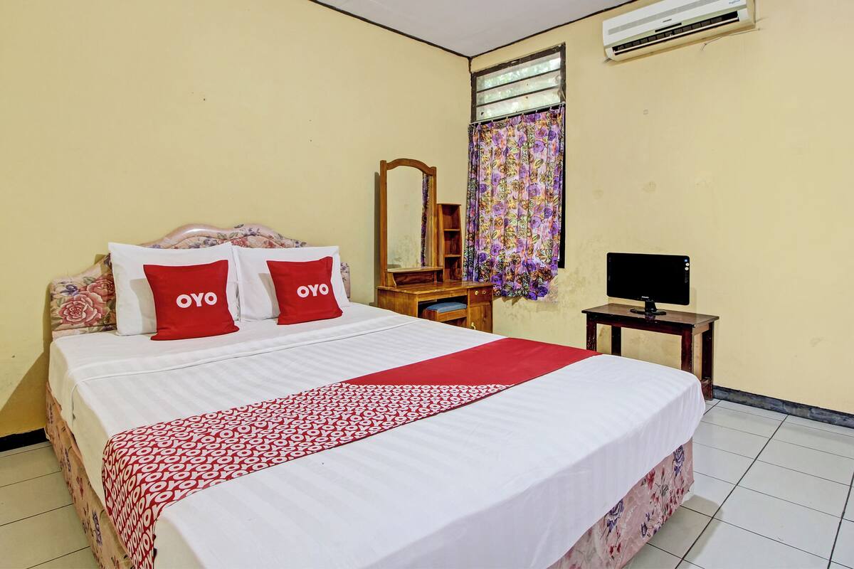 A neatly made bed with a patterned bedspread and two red pillows, a wooden nightstand with a television, a mirror, and a window with floral curtains.