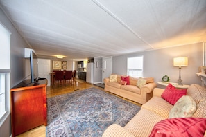 A spacious living room with a large sofa, a coffee table, and a patterned rug. There is a kitchen area in the background with a dining table and chairs.