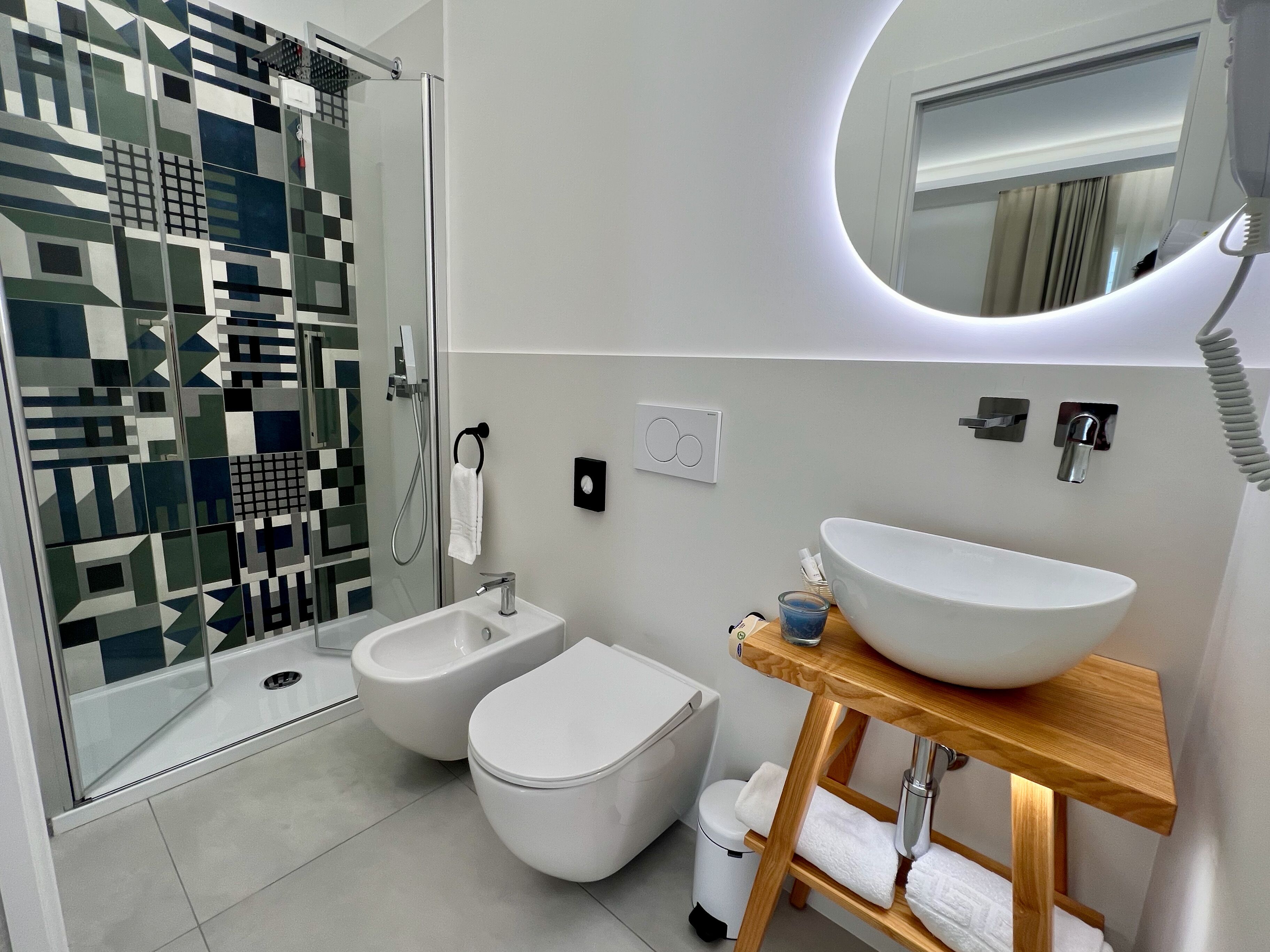 Une salle de bain moderne dotée d’une douche à parois de verre, d’un bidet, de toilettes, d’un grand lavabo avec un meuble-lavabo en bois et d’un miroir avec éclairage intégré.
