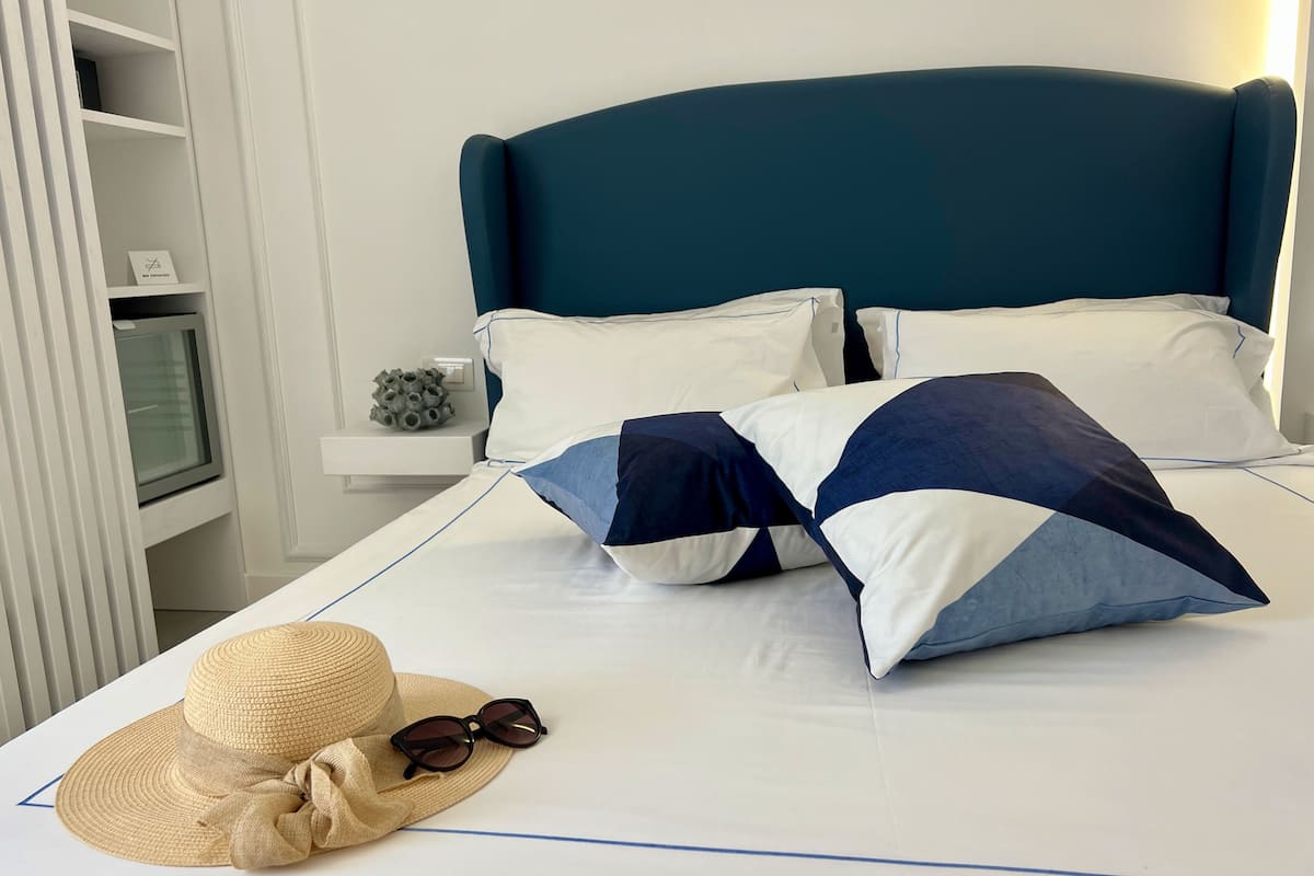 A neatly made bed with a teal headboard, white bedding, and blue and white pillows. A straw hat and sunglasses are placed on the bed.