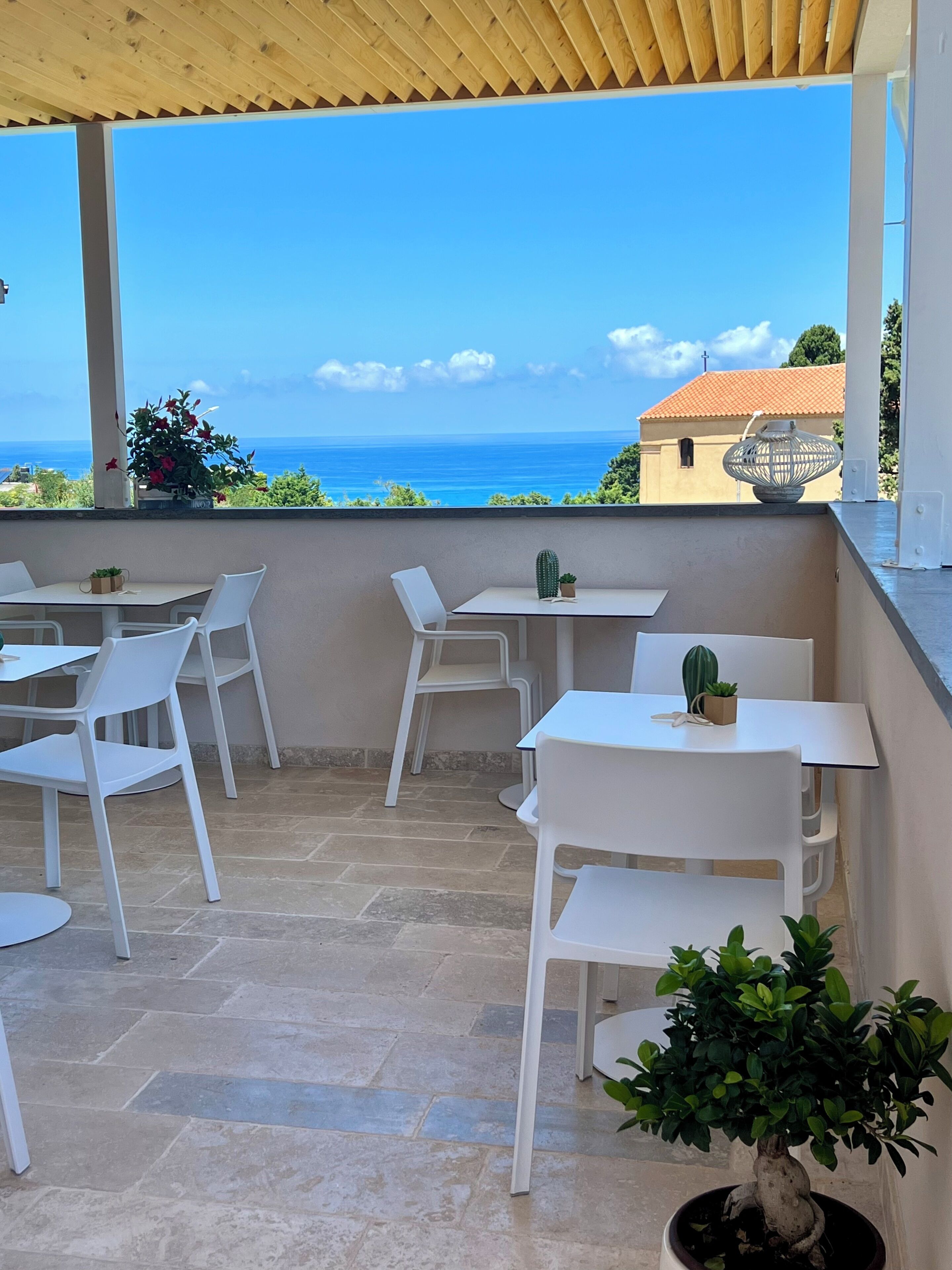 An outdoor seating area with white tables and chairs, a potted plant, and a view of the sea.