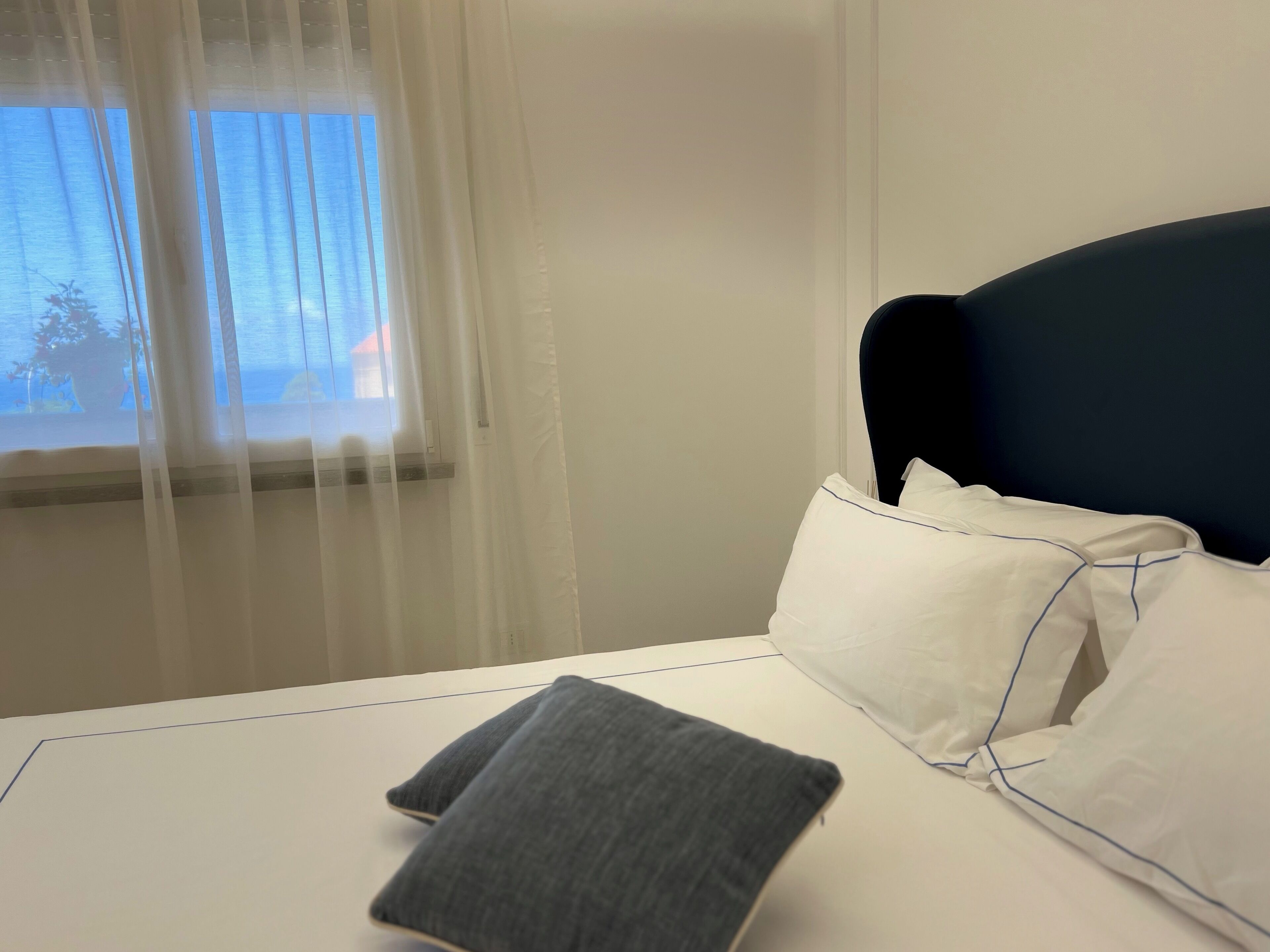 A hotel room with a bed, white pillows, a grey pillow, a window with sheer curtains, and a view of the outside.