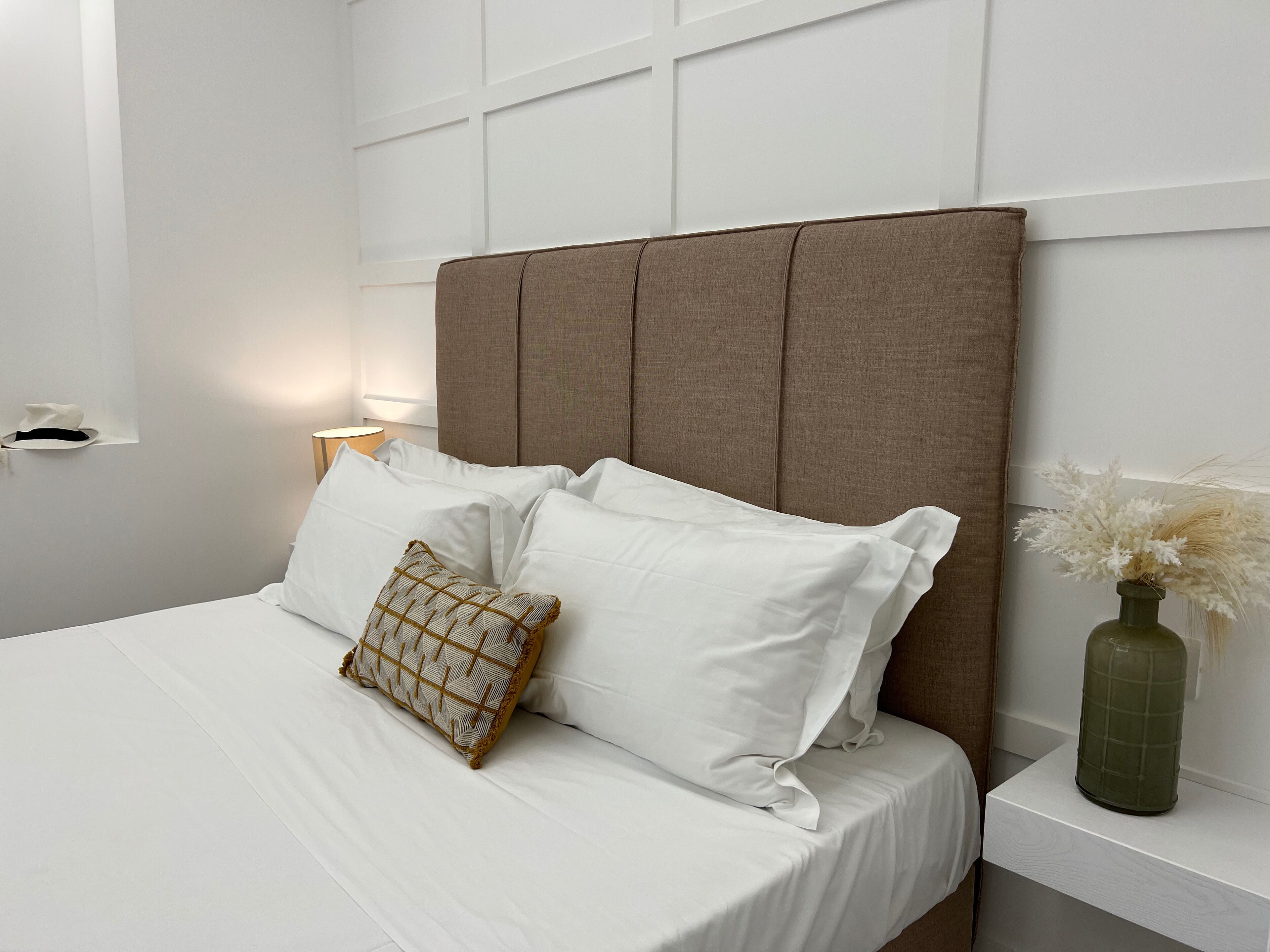 A neatly made bed with white linens and a patterned pillow, a brown upholstered headboard, a bedside table with a lamp, and a vase with dried flowers.