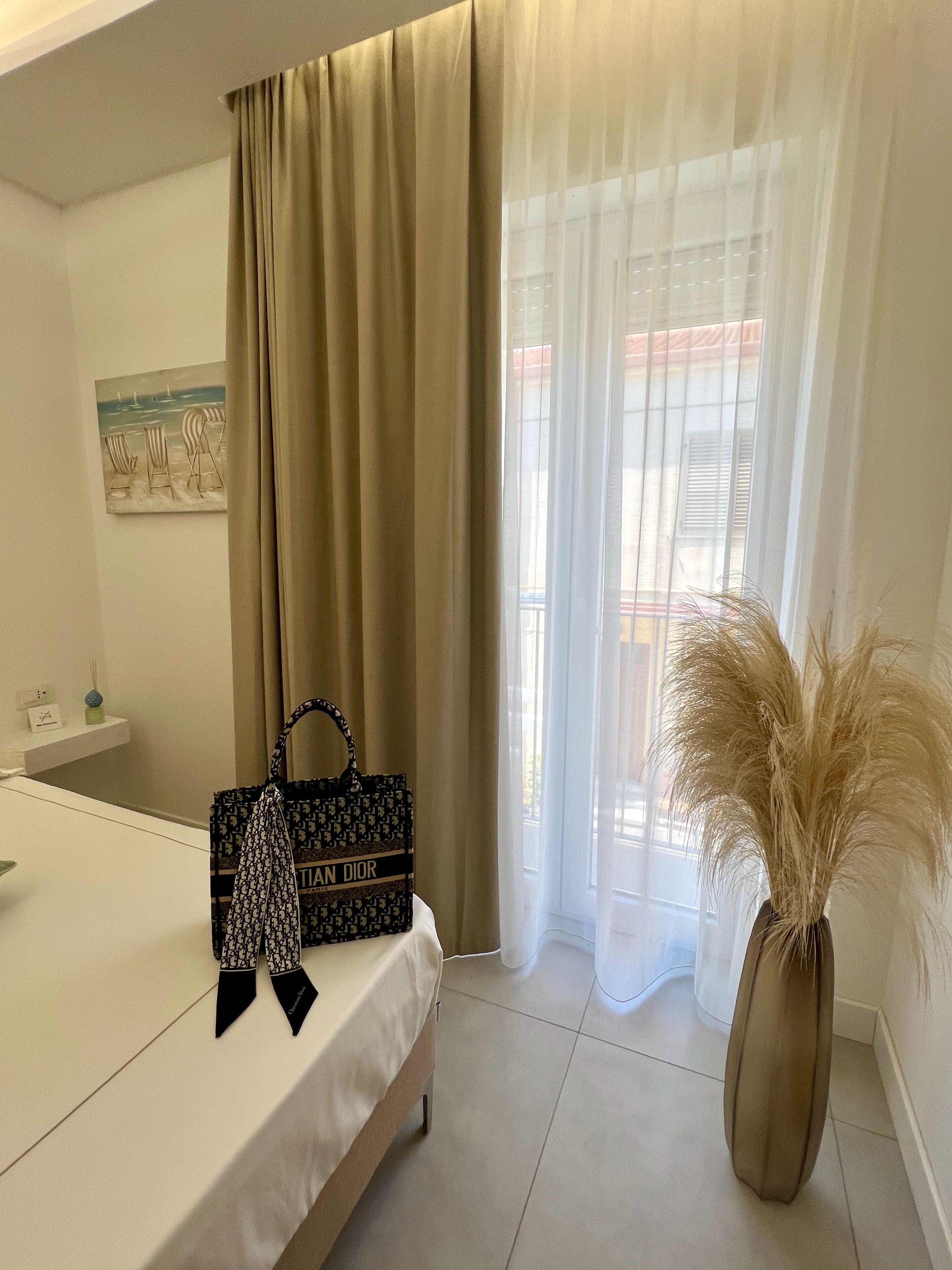Ein Hotelzimmer mit einem Bett, einem Schminktisch mit Spiegel, einem Gemälde einer Strandansicht, einer Handtasche und einer Vase mit Pampasgras.