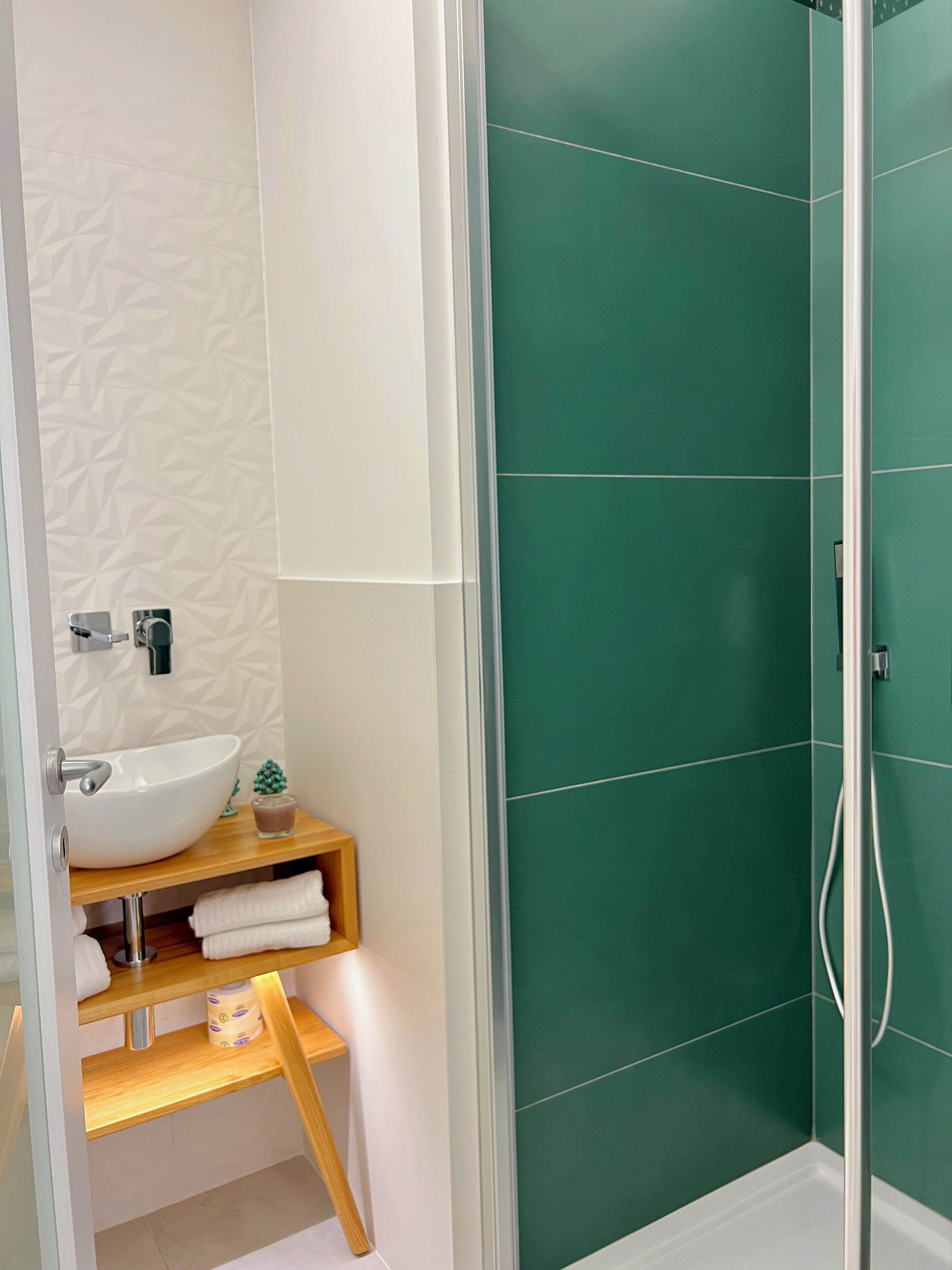 A bathroom with a white sink, green tiles, and a wooden shelf with towels.