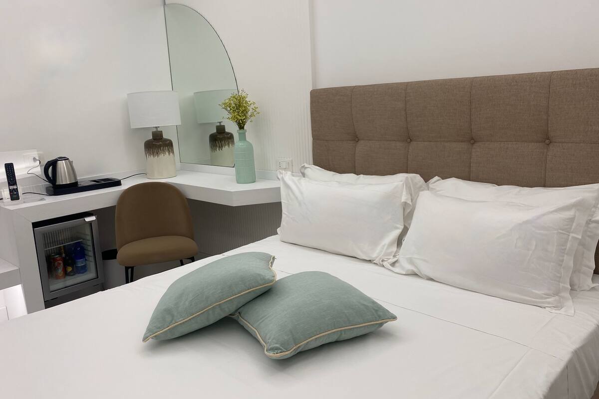 A hotel room with a bed, a desk, a chair, a mirror, and a minibar.